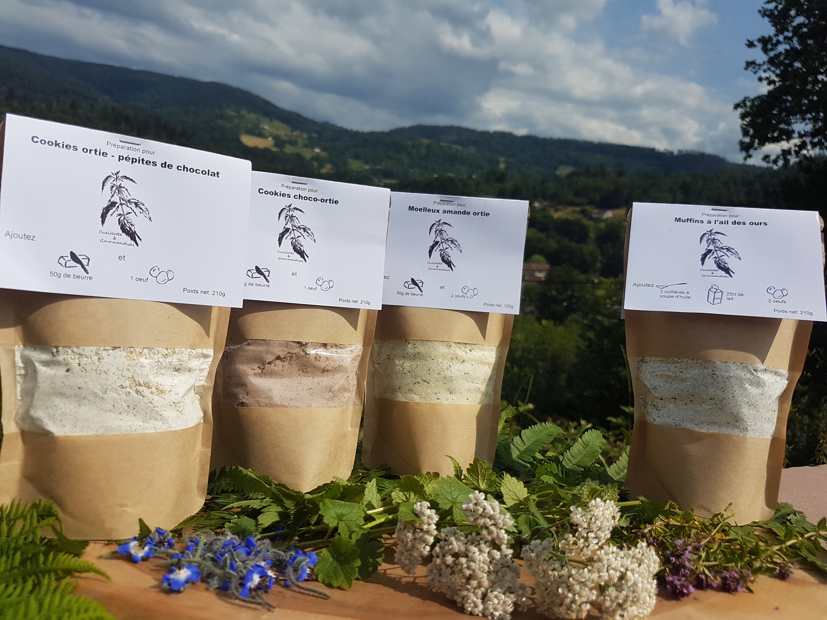Paquets de préparations de biscuits à base de plantes de Cueillette & Gourmandises (88290 Thiéfosse) devant un paysage vosgien

